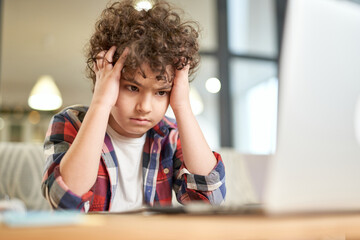 Tired kid. Portrait of tired latin boy holding his head, looking sad while studying at home using...