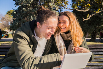 Young smiling couple sitting on a park bench consulting a laptop