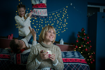 Mom and daughters are at home in winter. Family have fun together in the bedroom in winter. Mom with a cup of tea.  Children fight pillows on the bed. Quarantine, lockdown