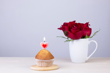 Red roses bouquet in white cup and little muffin cake with heart shaped candles on wooden background. Valentine day and holiday gift card with copy space