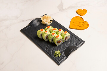 Side view of Japanese sushi roll with avocado slices and red caviar on top served with wasabi and ginger on black plate, heart shapes of turmeric powder spices on white marble background. Salmon roll
