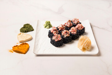 Side view of Japanese black rice sushi roll with unagi eel served with ginger, wasabi on square plate. Three Heart silhouettes of basil, garlic, turmeric powder spices on white marble background
