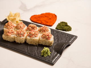 Close up to sushi roll with roasted salmon and tobiko flying fish roe on top served on black plate with wasabi and ginger. hearts of turmeric and basil spices and rose petals on marble background
