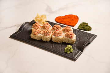 Side view of sushi roll with roasted salmon and tobiko flying fish roe on top served on black plate with wasabi and ginger. hearts of turmeric and basil spices and rose petals on marble background
