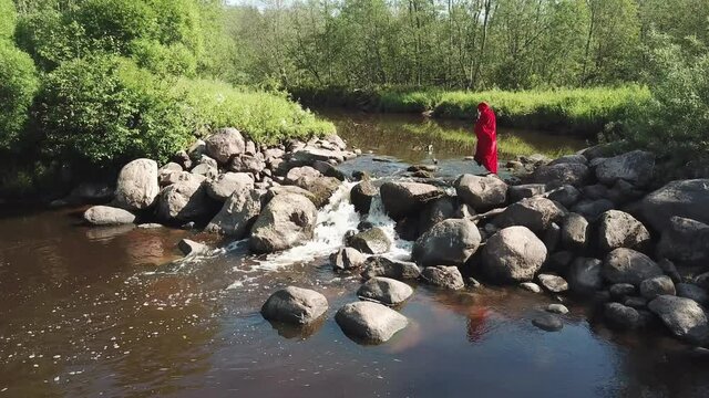 A girl in a red cloak crosses a stormy river with a waterfall, barefoot, without shoes, over the stones. Summer. Forest. View from above. Aerial photography. Shooting from a quadrocopter, drone.