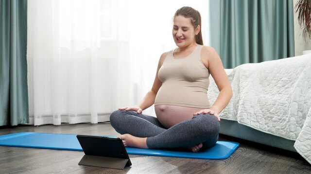 Happy smiling pregnant woman having online yoga lesson via video conference on tablet. Concept of healthy lifestyle, healthcare and sports during pregnancy