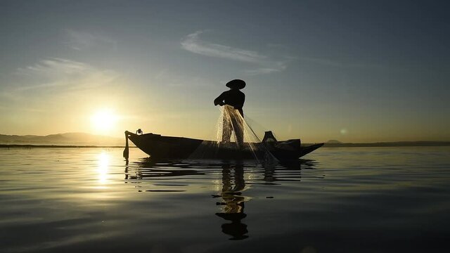 Video of silhouette scene of Asian fisherman stay on a boat throwing fishing net caught fish for food in a river or lake with beautiful sunrise sky