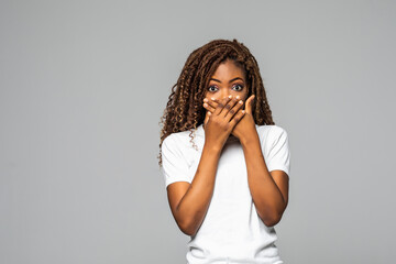 Shocked young African woman covering mouth with hands and looking at camera while standing against white background
