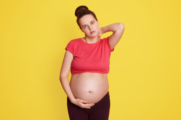 Pregnant woman standing with pain in neck, keeping hand on her bare tummy, looking directly at camera, has hair bun, wearing red t shirt, feels pain, being tired.