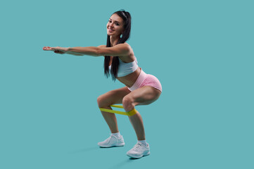 Young and slim smiling woman in sports clothing exercising with a resistance band against blue...