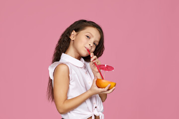 cute little child girl holding fresh juicy grapefruit and drinks juice isolated on pink background