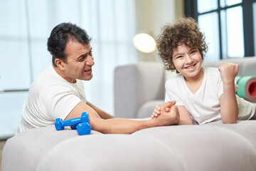 Fototapeta premium Strong guy. Happy latin boy looking satisfied after armwrestling together with his father at home, lying on the couch