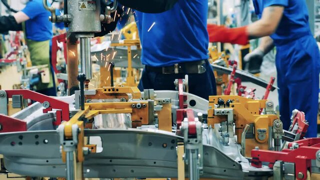 Metal automotive elements are getting spot welded by workers at modern industrial factory.