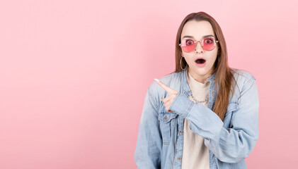 Horizontal picture of young female isolated on pink background wearing red eyeglasses, pointing leftwards with forefinger with surprise.