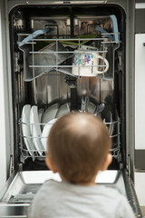 
Little child boy toddler helping mom to disassemble the dishwasher. The dishwasher is open, the child is sitting next to him and holding clean dishes in his hands.