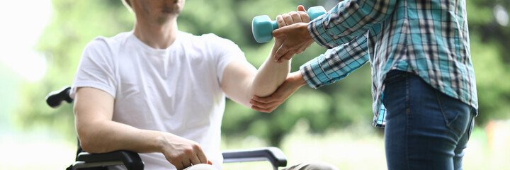 Close-up of middle-aged man sitting in wheelchair and trying to exercise with blue dumbbell. Woman helping male training arm. Sport and active lifestyle. Disabled people concept