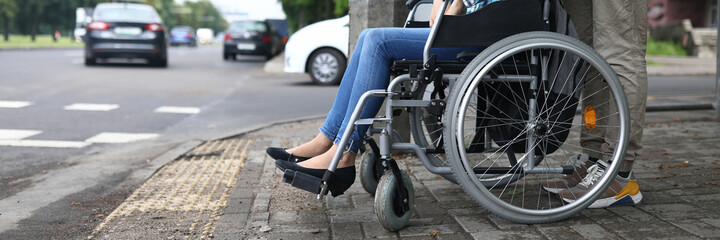Obraz premium Close-up of man accompany female in wheelchair. Person taking care of lover. Carefully crossing street. Lady in jeans and blue shirt. Walk in city. Disabled people concept