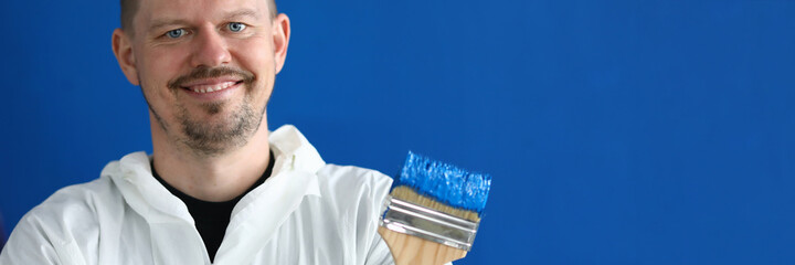 Fototapeta premium Portrait of paintbrush covered with blue paint. Smiling male worker in uniform on wall background. Professional interior designer. Renovation and construction site concept