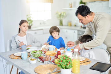Caring latin father serving his adorable kids while having breakfast together at home