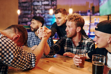 Friends plays arm wrestling. Group of people together indoors in the pub have fun at weekend time