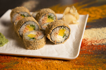 Close up of Japanese Tempura hot sushi roll with roasted salmon served with wasabi, ginger on white square plate. Dark background with scattered spices: dried garlic, turmeric, curry, paprika powder
