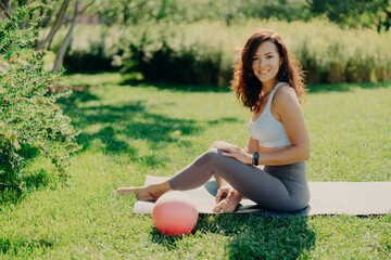 Positive brunette woman in cropped top and leggings poses bare feet on karemat uses ball for...