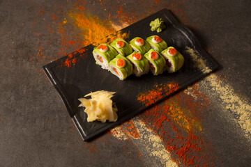 Top view of Japanese sushi roll with avocado and red caviar on top served with wasabi and ginger on black plate. turmeric powder, dry garlic , paprika spices on dark background. Salmon inside roll

