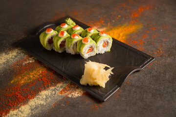 Japanese sushi roll with avocado and red caviar on top served with wasabi and ginger on black plate. turmeric powder, dry garlic, paprika spices on dark background. Salmon wrapped inside roll
