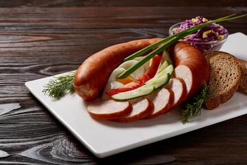 Sausages with a garnish of vegetables and herbs with sauces and bread on a white rectangular plate on a dark wooden table.