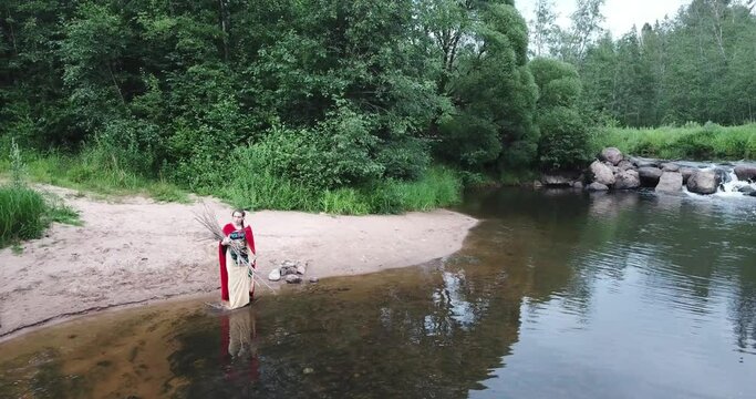 girl crosses river into a ford, carries sticks, branches, firewood in her hands. Walking over stones along river bank with waterfall. sandy beach. A fabulous image, fabulous character in a red cloak.