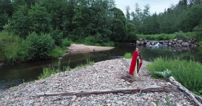 girl in a red cloak carries firewood and brushwood in her arms. puts firewood into a fire. Preparing to make a fire, a bonfire. river flows, a waterfall. Beautiful nature. Fairy image, fairy character