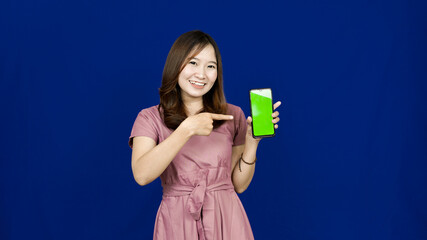 beautiful asian woman pointing green screen in handphone isolated blue background