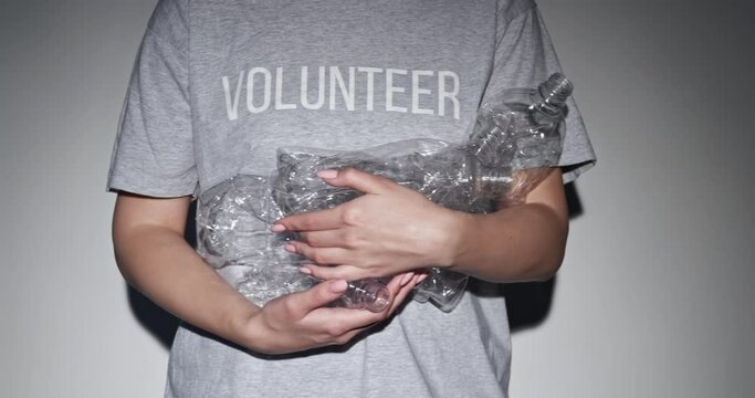 Plastic reduction. Zero waste. Nature conservation. Unrecognizable volunteer woman in t-shirt holding used creased bottles pile in spotlight on gray background.
