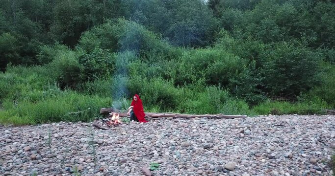 girl in a red cloak sits near the fire. Looks around. Fabulous image, fairytale character. Flying on a quadrocopter, drone from parva to the left.