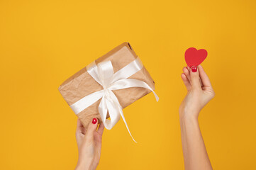 Female hands holding a paper heart and gift box on yellow background. Concept of a gift for the holidays, birthday, Christmas, wedding. Flat lay, top view