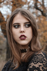 The face of a young witch. Makeup in the Gothic style. Spooky atmosphere in the autumn garden. Brunette with painted lips. The gaze is set aside. Black clothes. Shooting for Halloween.