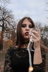 Girl holding a glass goblet with a garland in the middle. Autumn shooting in the park. Gothic photo shoot. Brunette dressed in a black dress and accessories.