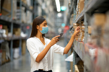 The life during outbreak of Covid-19 concept: Young Asian female with face mask use mobile phone check model of goods on carton in department store.