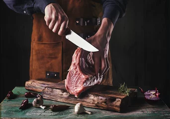 Fotobehang A guy in a leather apron is slicing raw meat. The butcher cuts the pork ribs. Meat with bone on a wooden cutting board.. © Сергей Опиканец