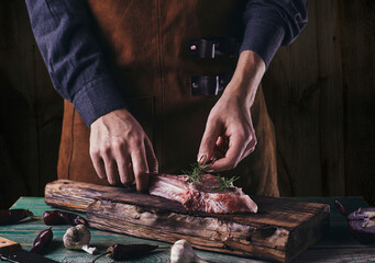A man in a leather apron lays a rosemary branch on a steak with a bone. Meat on a wooden cutting board..