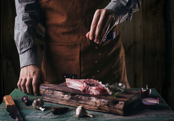 A guy in a leather apron sprinkles black pepper on a steak with a bone. Raw meat on a wooden cutting board..
