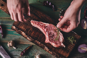 Pork steak with bone on a wooden cutting board. Man's hands lay a rosemary branch on a piece of meat..