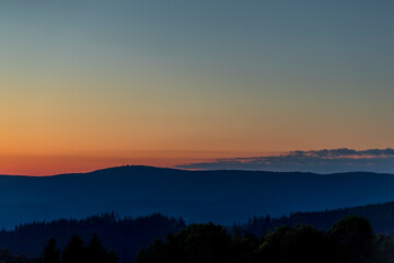Sun setting over a horizon of orange hills with a coniferous tree on the left and clouds moving in the background over the Beskydy countryside.