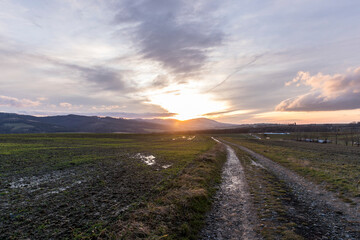 A dirt road leading around a field during an orange sunset on the horizon of the setting sun behind the hills.