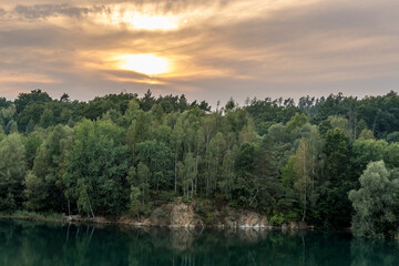 Fototapeta premium Flooded quarry and trees lying on the shore during sunset with fast moving clouds and a blue-orange sky in the background.