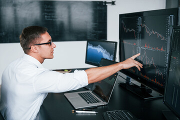 Young male stock brocker works indoors in the office. Many of displays