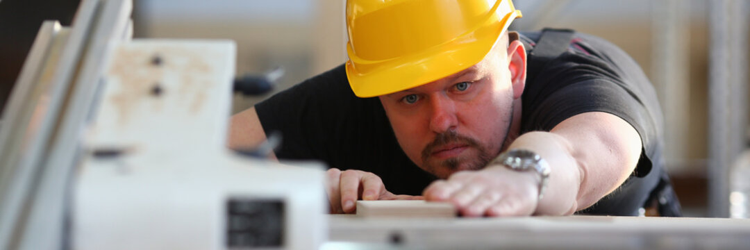 Young Bearded Carpenter In Hard Hat Is Working With Wooden Board And Using Electric Equipment