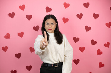 Young beautiful woman over pink background with harts showing and pointing up with fingers number one while is serious