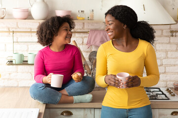 Happy Black Mother And Little Daughter Talking And Eating Snacks In Kitchen