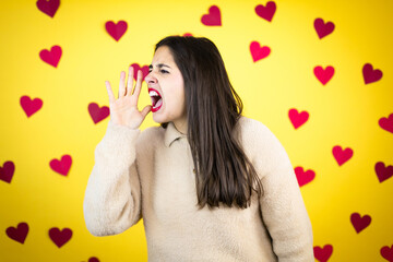 Young caucasian woman over yellow background with red hearts shouting and screaming loud to side with hand on mouth